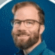 Smiling man with glasses and a beard, wearing a blue shirt, against a blue background.
