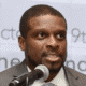 Man speaking at a microphone in front of a blurred background with text.