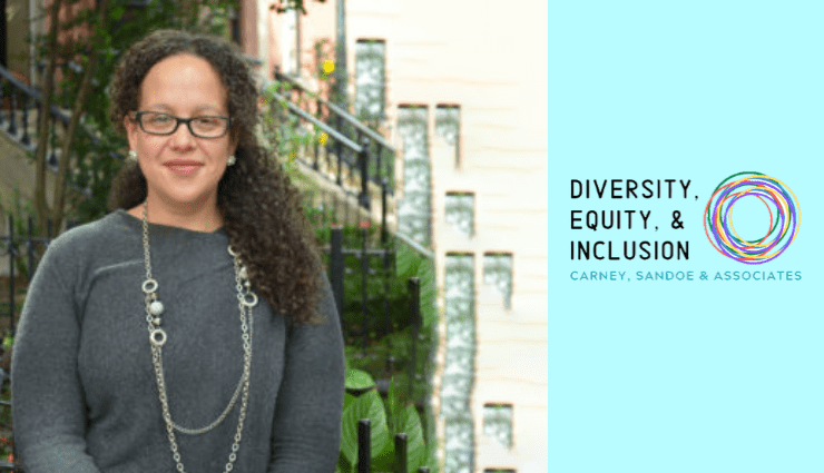 Person standing outdoors wearing glasses, with braided hair and a necklace, next to a wall with diversity and inclusion text.