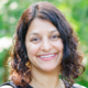 Smiling person with curly hair standing outdoors with a green, blurred background.