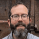 Man with glasses and a beard smiling in front of a wooden background.