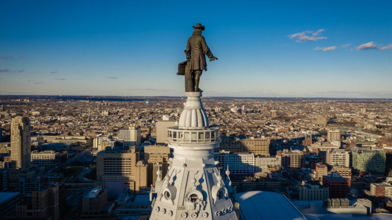 William Penn statue atop Philadelphia City Hall with cityscape in the background under a clear blue sky.
