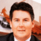 A person with dark hair and a suit, blurred background with abstract art and architectural elements.