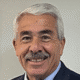 Smiling older man with gray hair and mustache wearing a dark suit against a plain background