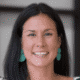 Smiling person with long dark hair and turquoise earrings, wearing a patterned top against a blurred background.