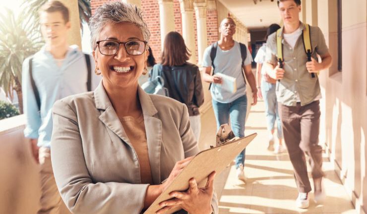 Smiling woman with clipboard standing in a corridor surrounded by students walking to class.