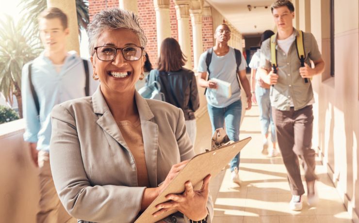 Smiling woman with clipboard standing in a corridor surrounded by students walking to class.