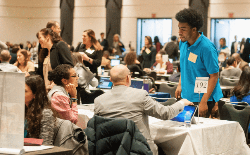 People networking at a busy career fair, with a young person talking to a professional at a table.