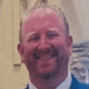 Man smiling in a suit against a blurred indoor background.