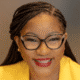 Smiling person with braided hair and glasses wearing a yellow jacket against a gray background.