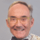 Smiling older man with glasses, wearing a patterned shirt against a plain background.