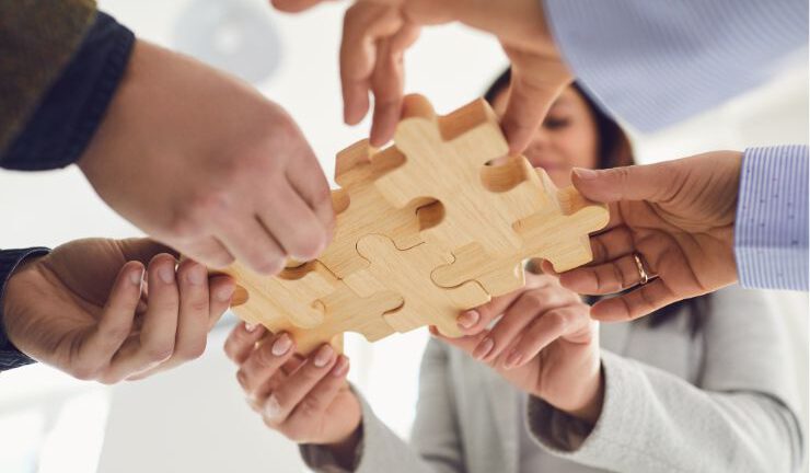 Hands of diverse people assembling a large wooden puzzle piece together.