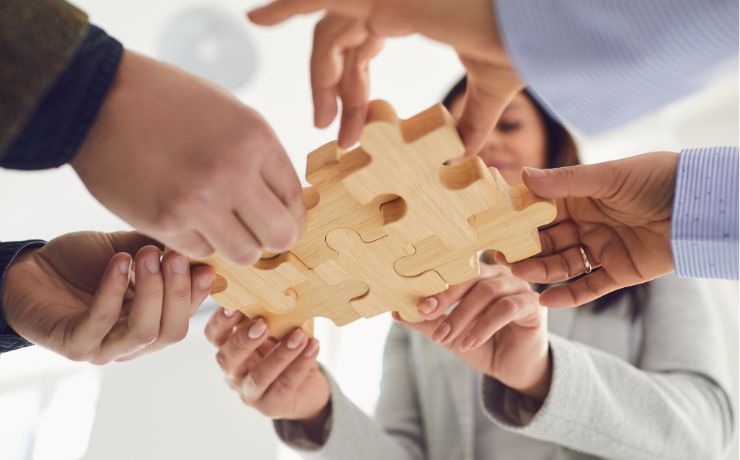 Hands of diverse people assembling a large wooden puzzle piece together.