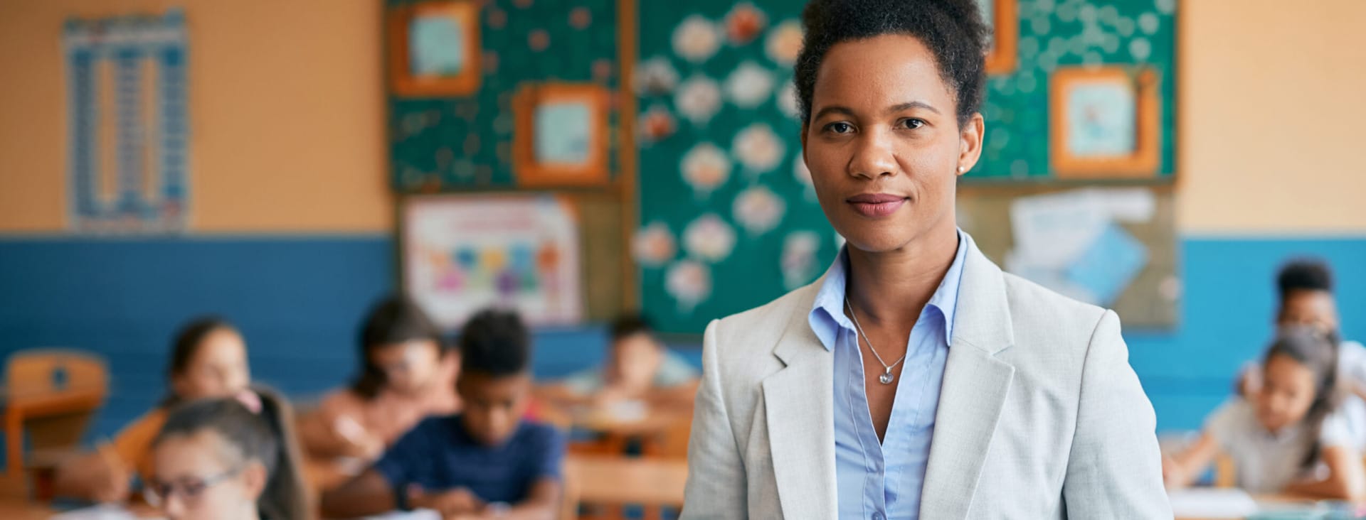 Teacher holding papers in a classroom with students working at desks in the background.