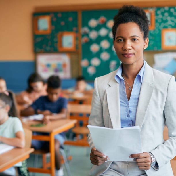 Teacher holding papers in a classroom with students working at desks in the background.