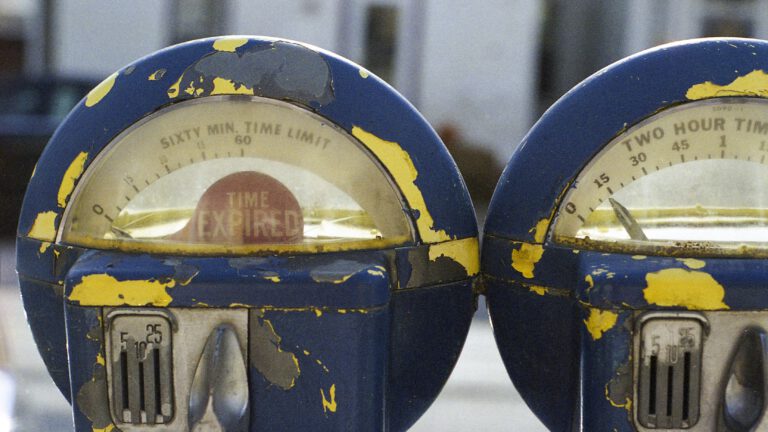 Two old, weathered parking meters with peeling blue paint and visible internal mechanisms, set against a blurry background.