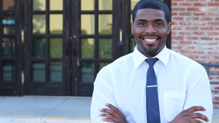 Male teacher with his arms crossed smiling in front of a school door outside