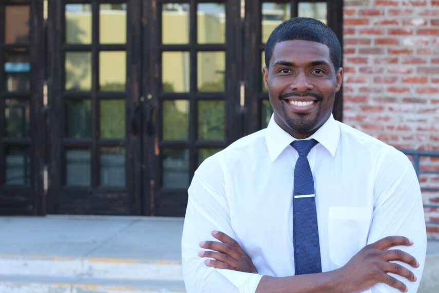 Male teacher with his arms crossed smiling in front of a school door outside