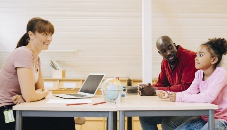 Three people sitting at a table with laptops and notepads, engaged in a discussion in a modern office setting.