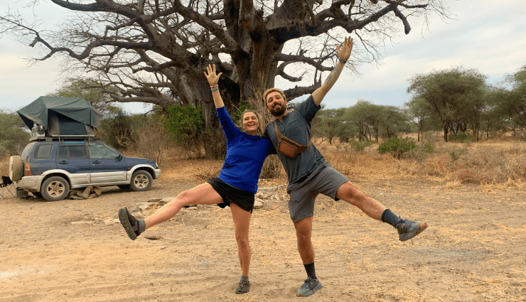 Two people joyfully posing with raised arms in front of a large tree in a dry landscape with a parked car nearby.