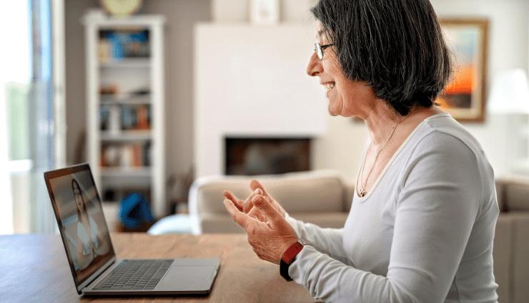 Older woman smiling during a video call on a laptop, sitting in a cozy living room with shelves in the background.
