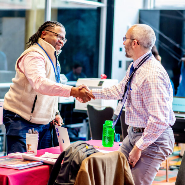 Two people shaking hands at a conference table, both smiling and wearing lanyards, with conference materials on the table.