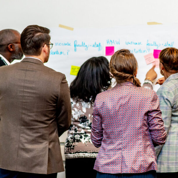 Group of people collaborating on a project, facing a whiteboard with colorful sticky notes, engaging in a discussion.