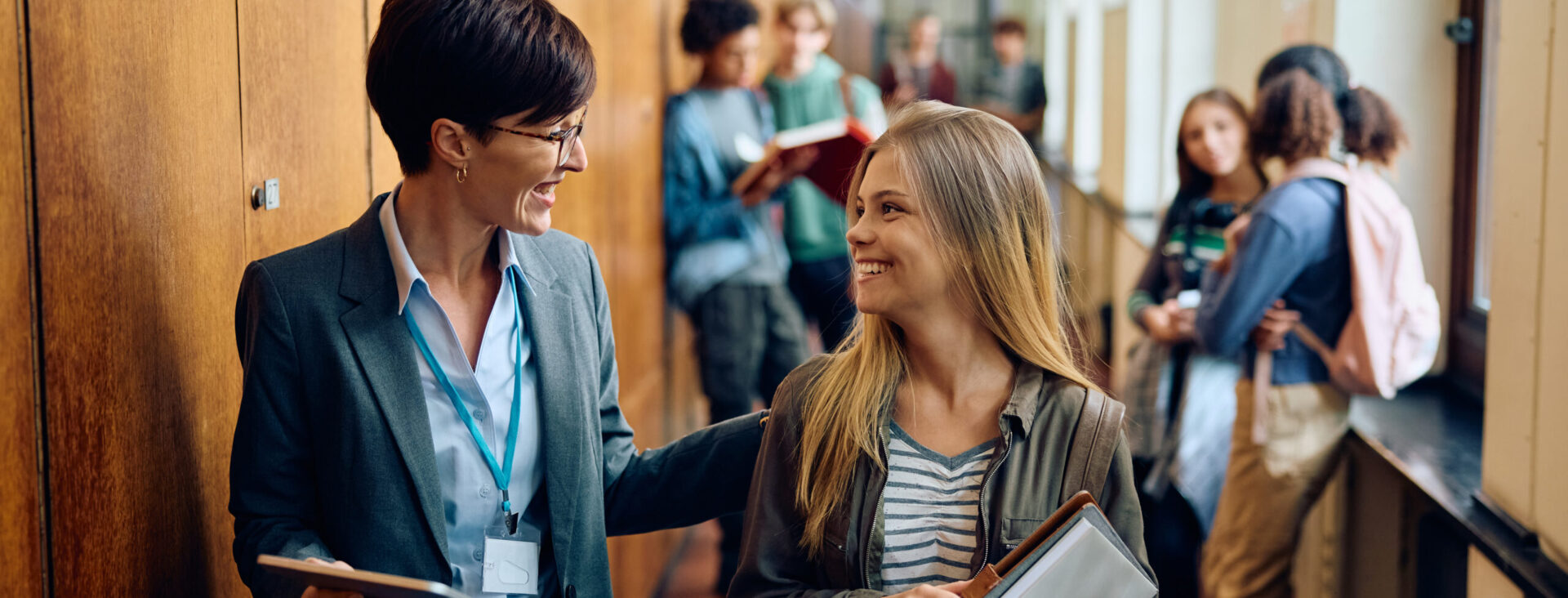 Teacher and student talking in a busy school hallway filled with students, some holding books and notebooks.