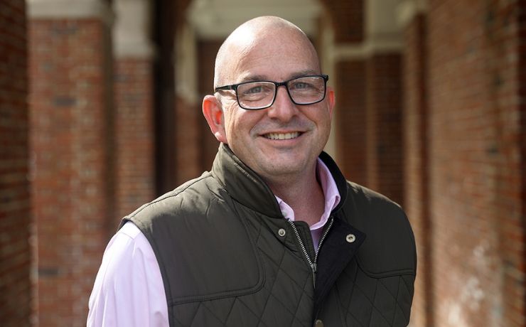 Bald man with glasses smiling, wearing a pink shirt and a dark vest, standing in front of a building with brick arches.