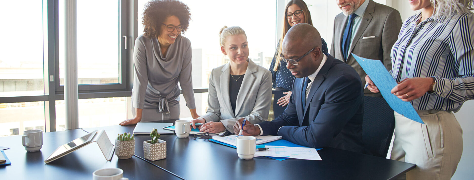 Diverse group of businesspeople standing around their manager sitting at an office boardroom table reviewing charts and paperwork