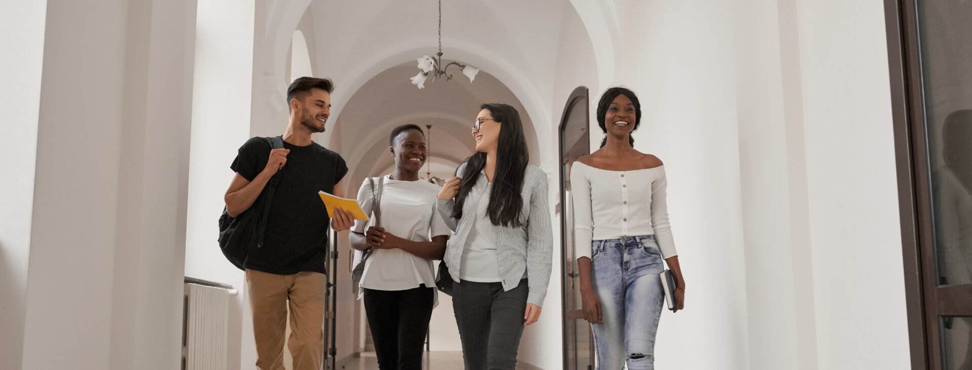 International group of four students with backpacks walking at university and resting after lecture. Happy friends of Caucasian and African studying together, spending free time on brake and smiling.