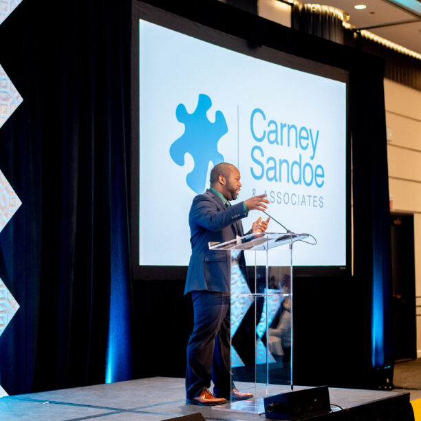 Speaker presenting on stage with Carney Sandoe and Associates logo displayed on a screen in the background