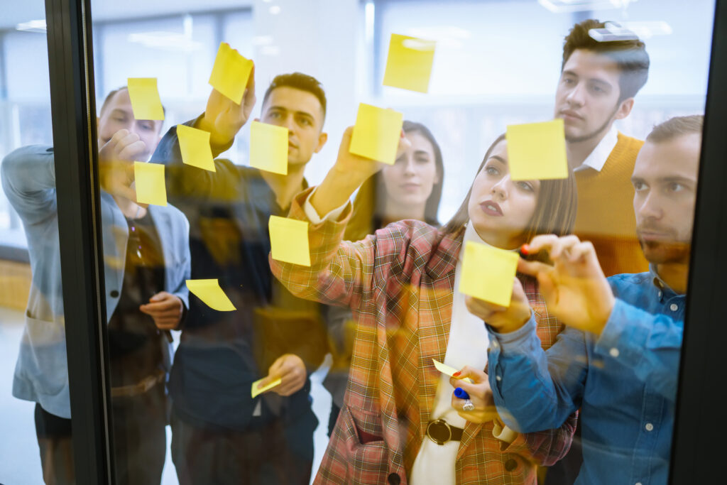Group of people brainstorming with sticky notes on glass wall in a bright, modern office environment.