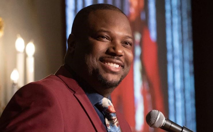 Smiling man in a suit speaking into a microphone at an indoor event.