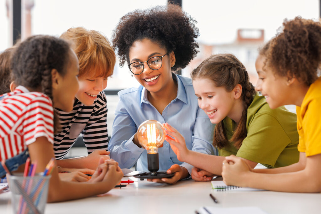 Multiethnic teacher and kids studying electricity and incandescent light bulb at physics lesson in bright classroom