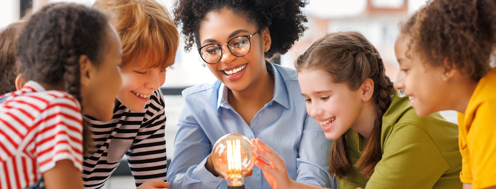 Multiethnic teacher and kids studying electricity and incandescent light bulb at physics lesson in bright classroom