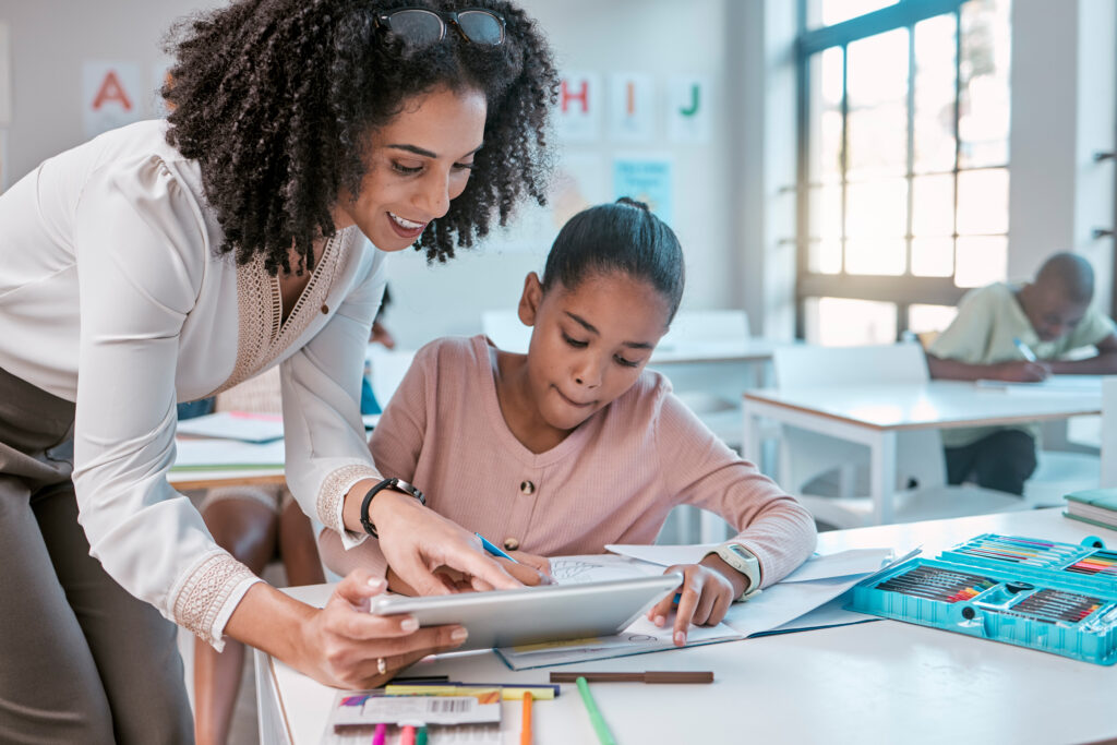 Female teacher helping young girl, teaching creative work in book and children in sunny classroom. Education empowerment in school, educator speaking to student and learning mathematics at desk.