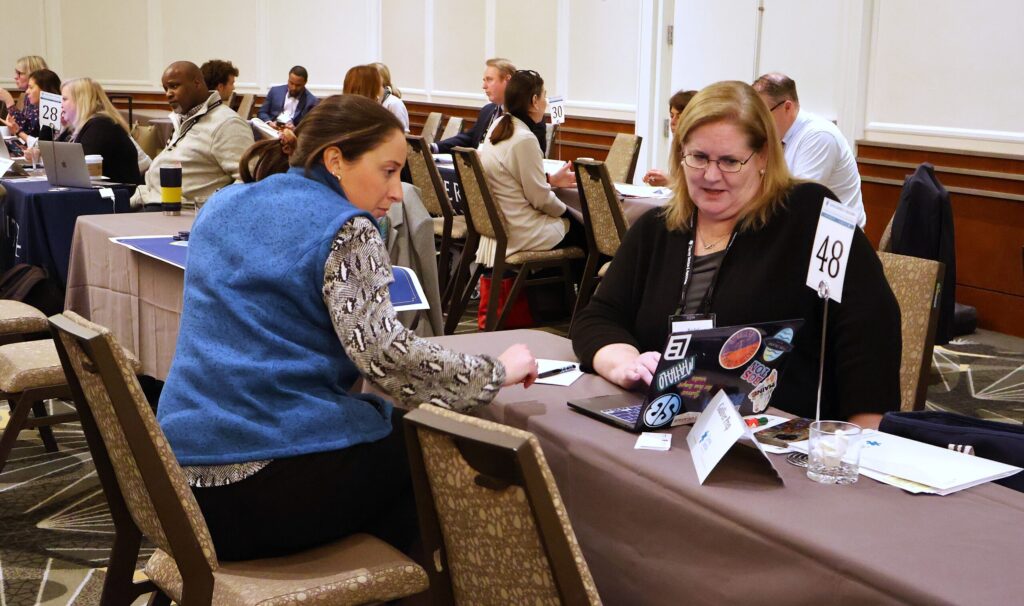 Two people sitting at a table during a business meeting, engaged in conversation with documents and laptops in front of them.