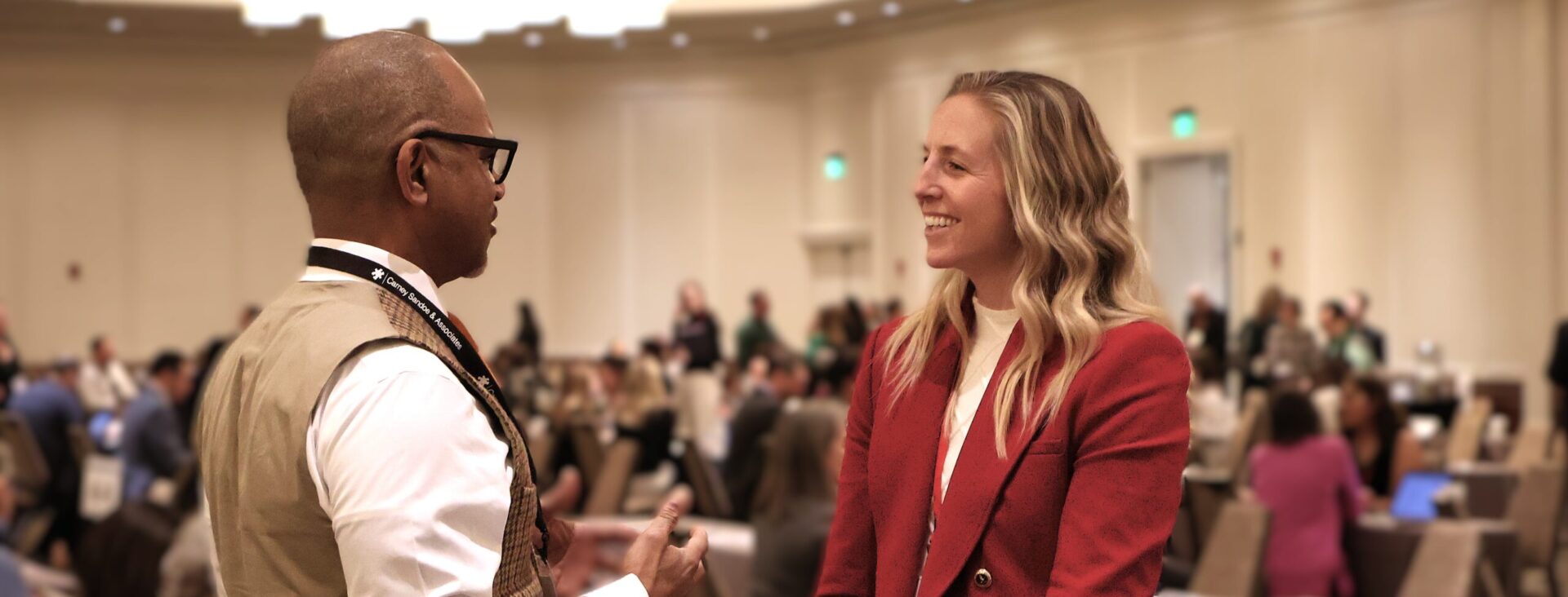 Two people engaged in conversation at a conference with a busy background.