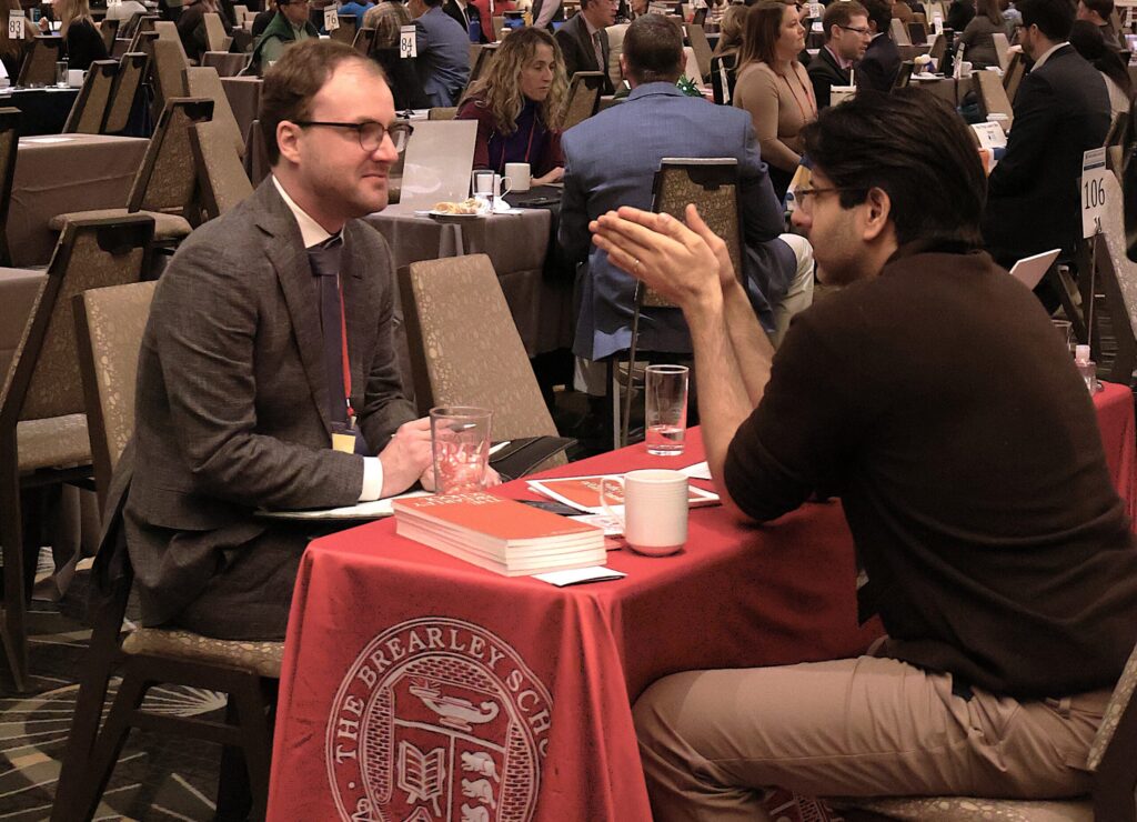 Two people having a discussion at a conference table with books and coffee cups in a crowded room.
