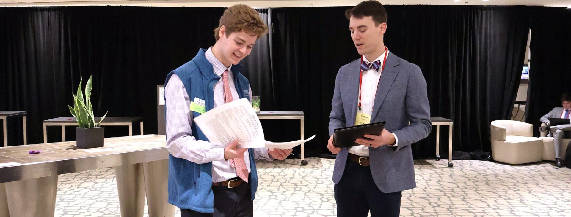 Two people in a room discussing documents, standing on a patterned carpet with tables and equipment in the background.