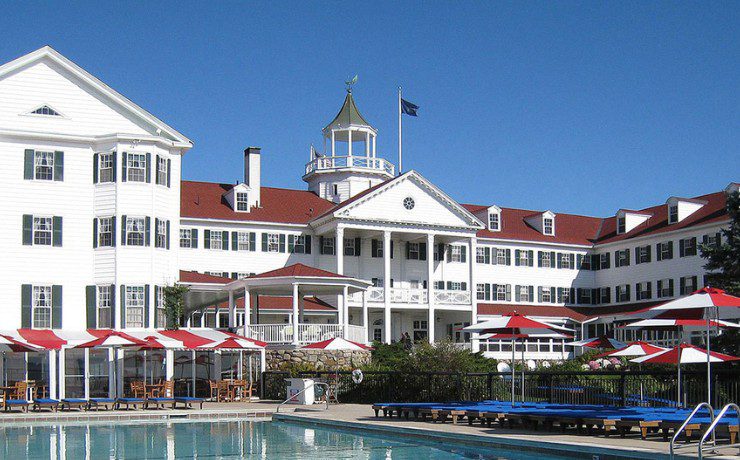 Historic hotel with a red roof, white facade, and a swimming pool in the foreground on a clear day.