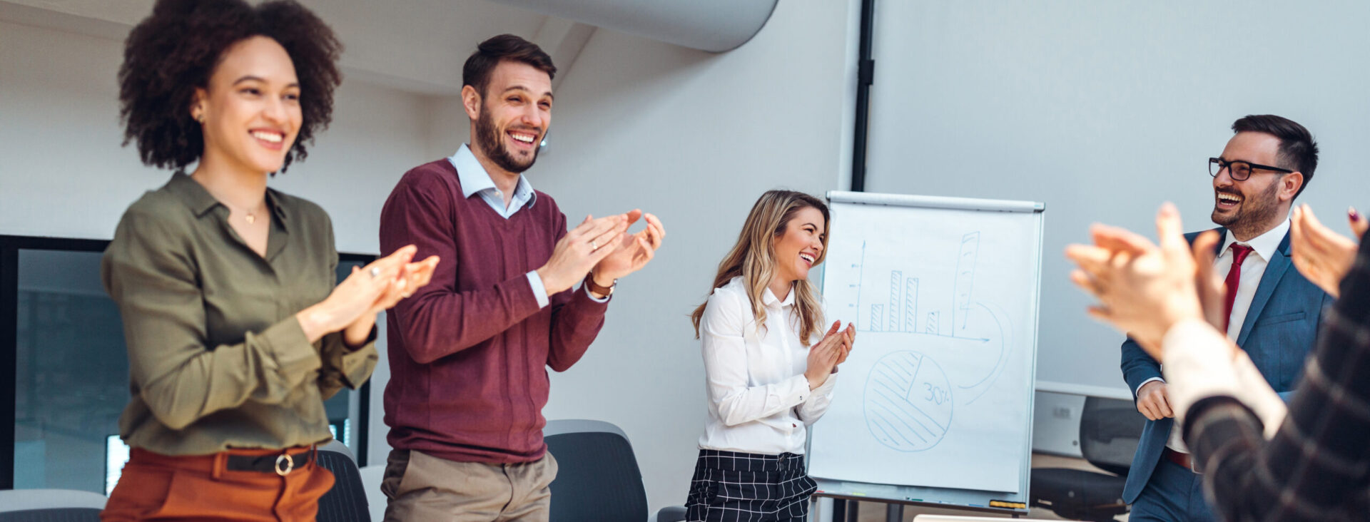 Group of colleagues standing around a table, clapping and smiling during a meeting in a modern office setting.