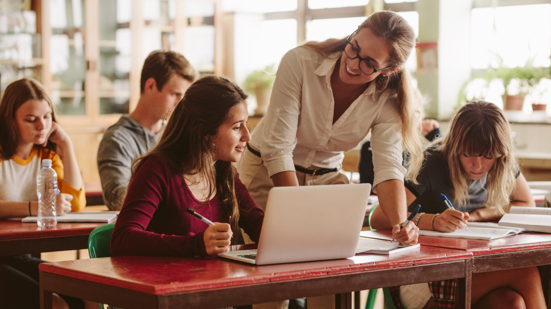 Teacher assisting a student with laptop in a classroom setting, surrounded by other students working at desks.