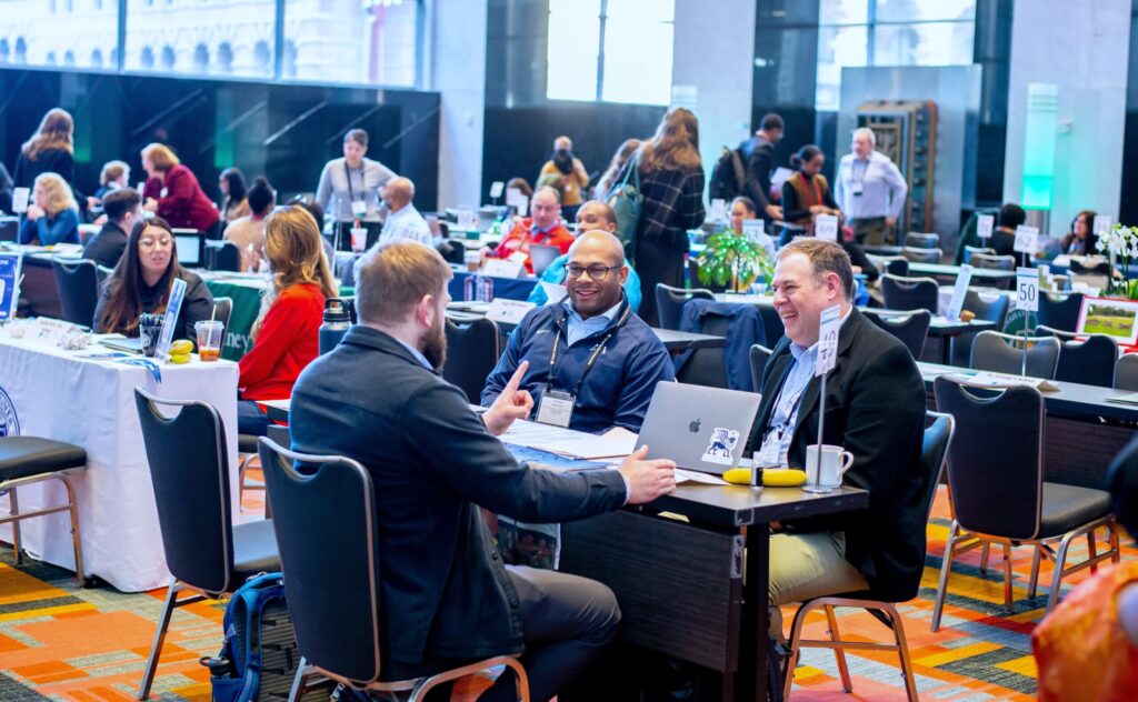 People at a conference table engaged in discussion, smiling and interacting in a busy conference hall.