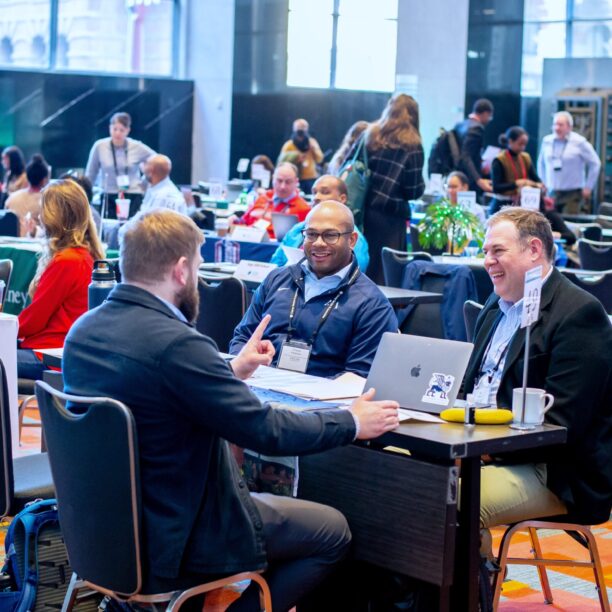 People at a conference table engaged in discussion, smiling and interacting in a busy conference hall.