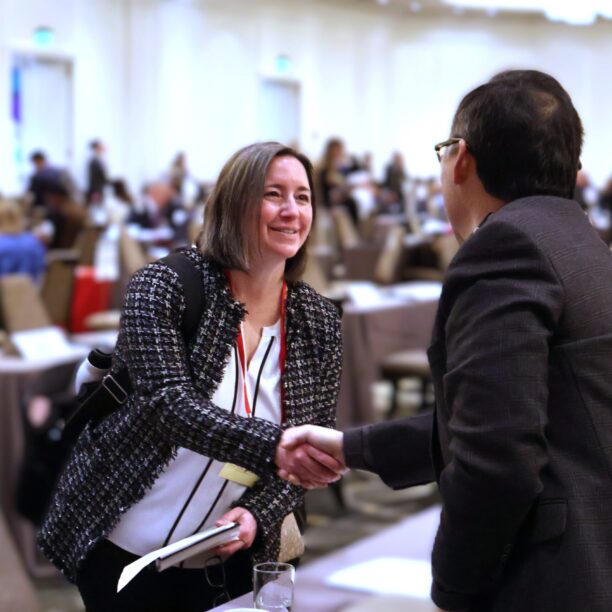 Two people shaking hands in a conference hall filled with attendees sitting at tables.