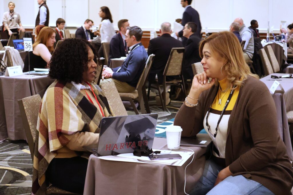 Two women sitting at a table, engaging in conversation at a busy conference setting with laptops and coffee cups.