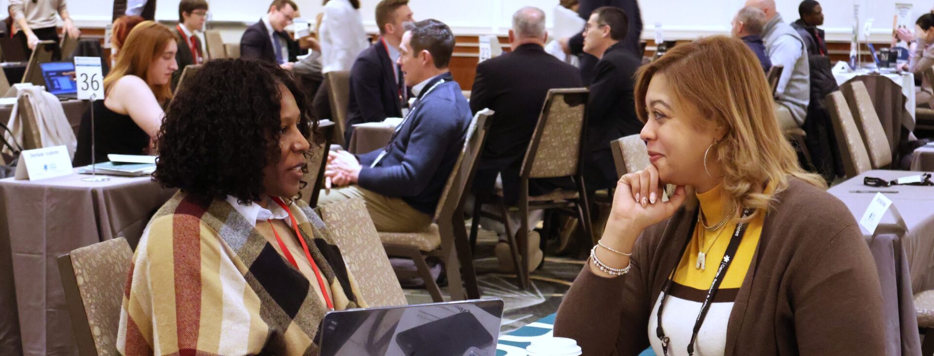 Two women sitting at a table, engaging in conversation at a busy conference setting with laptops and coffee cups.