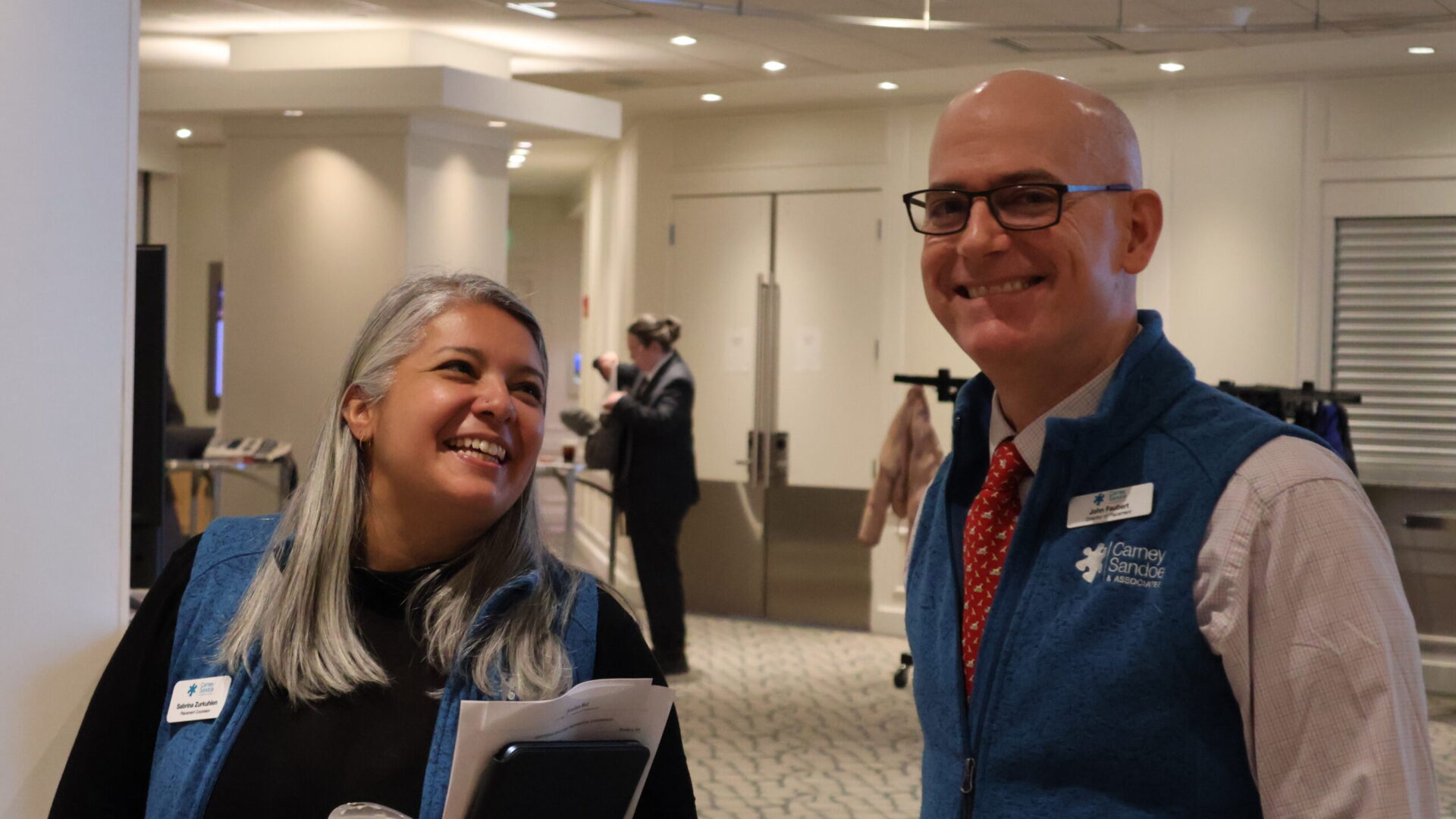 Two people smiling and conversing in a professional setting, one holding a drink and the other wearing a name badge.